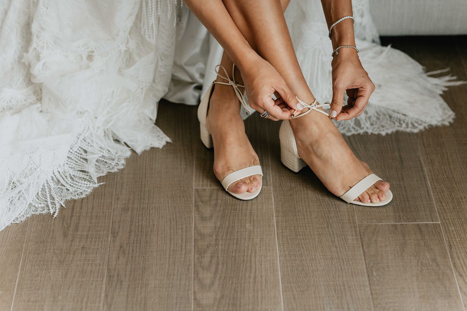 Bride fastening delicate white sandals with lace wedding dress hem visible.
