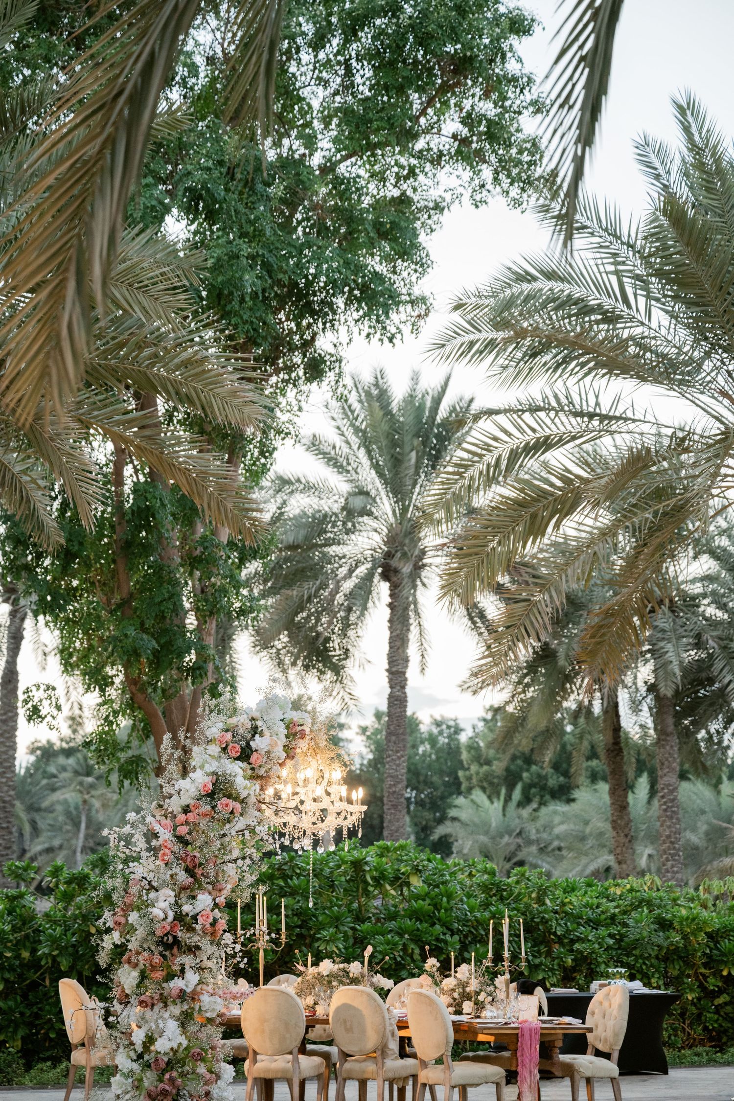 Romantic outdoor dining setup under palm trees with crystal chandelier and floral arrangements.