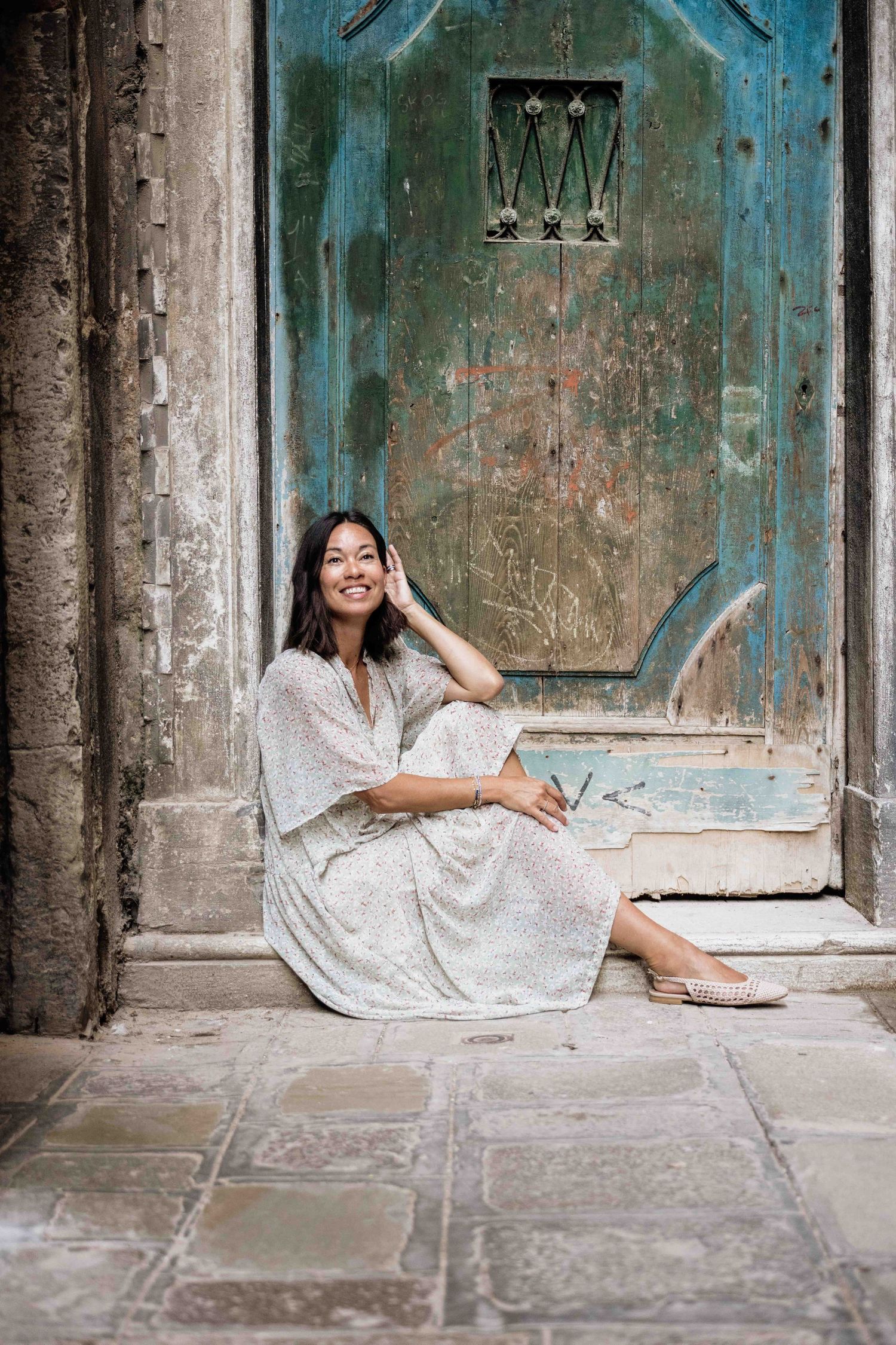 Person in white flowy dress poses elegantly against weathered turquoise door in Venice.