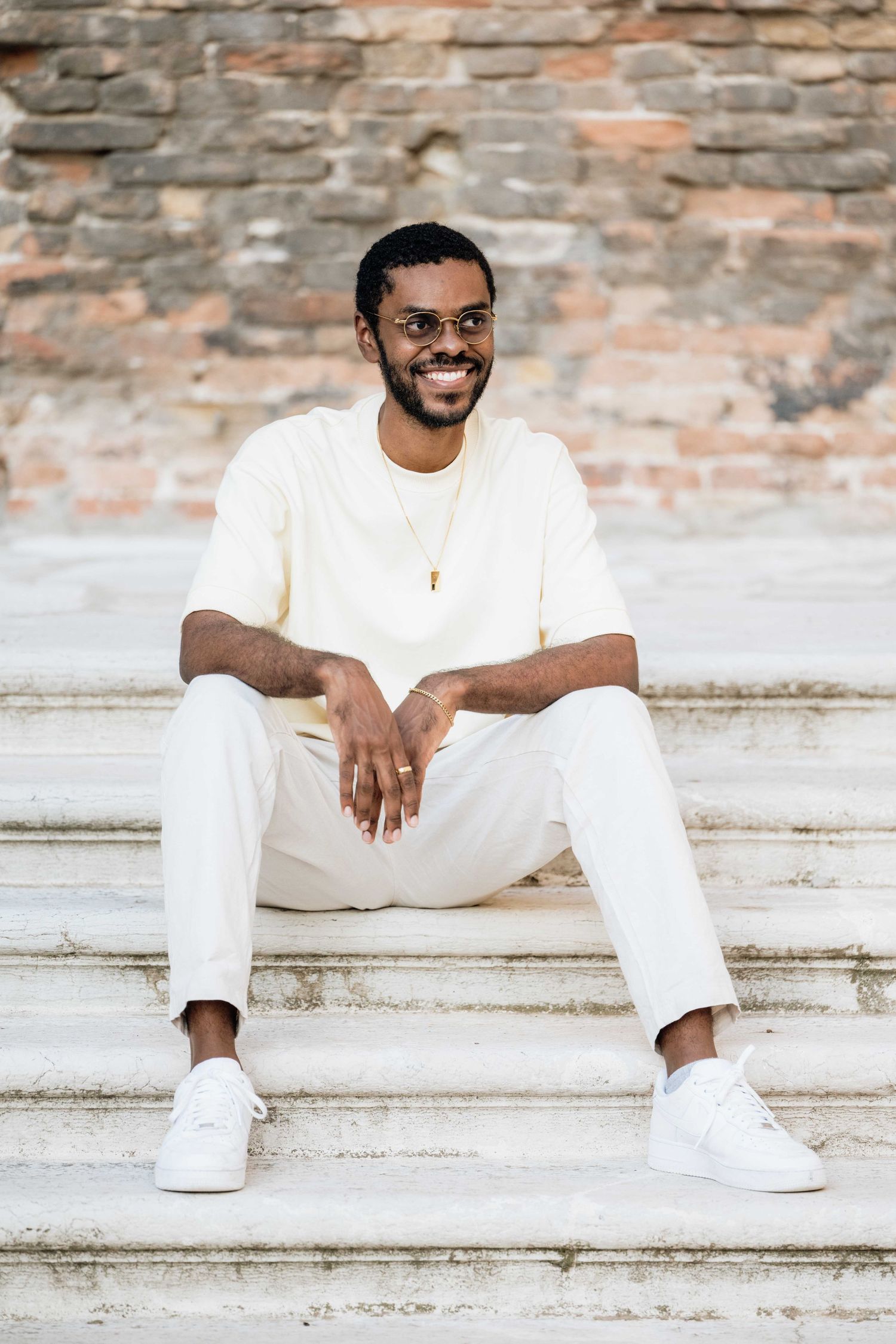 Person in all-white outfit sits casually on concrete steps near brick wall.