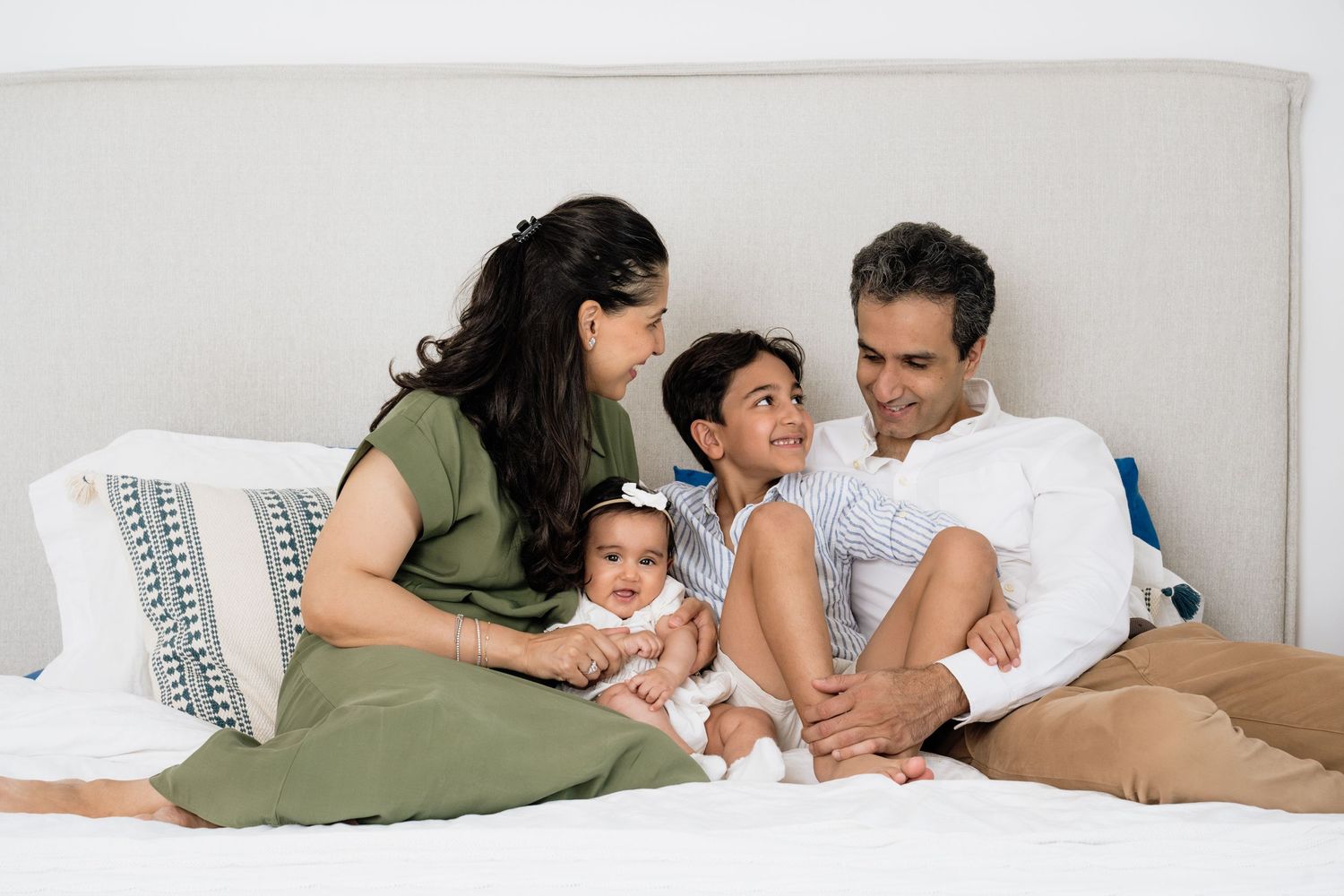 Family lounging together on white bedding in casual home setting.