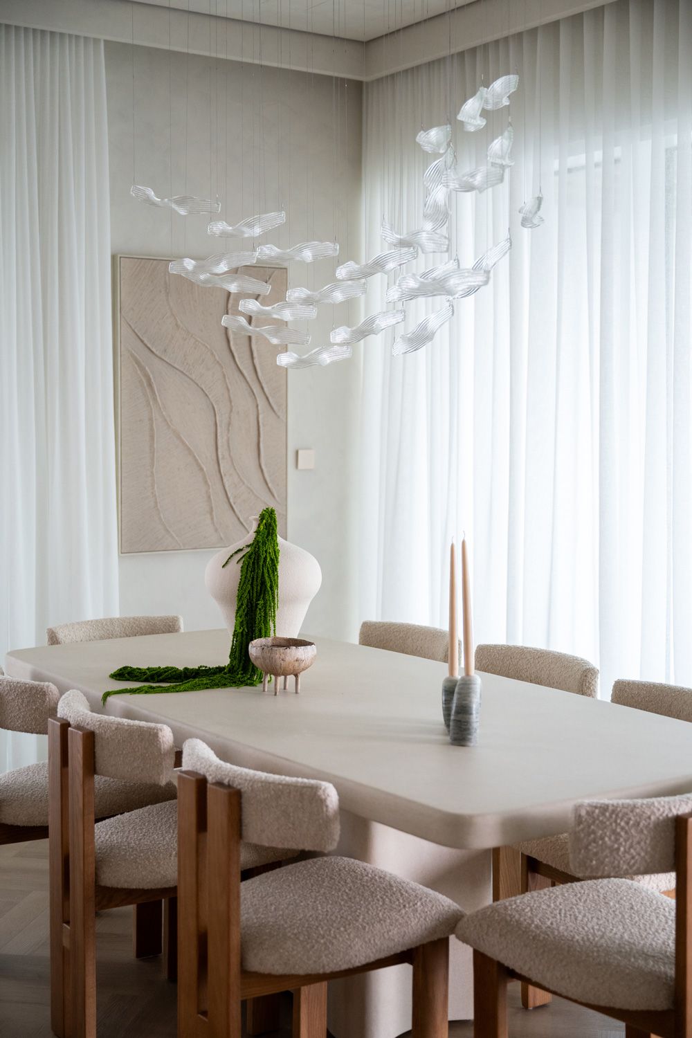 Modern dining room with beige upholstered chairs, white table and delicate branch-like lighting fixture mounted on wall.
