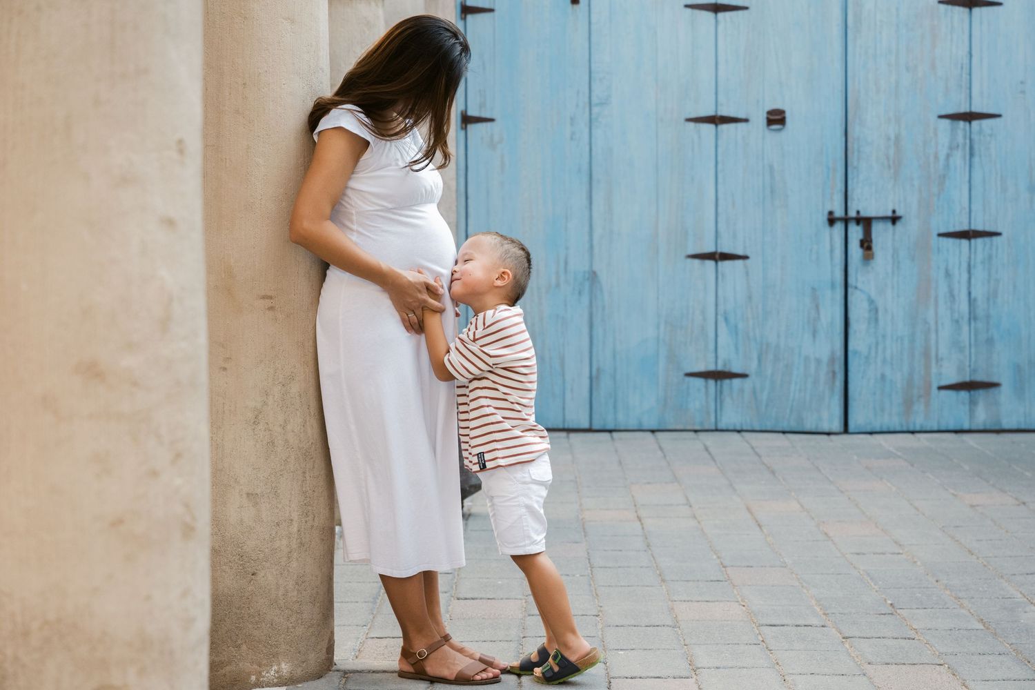 Mother and child in white outfits stand against blue rustic doors in stone alleyway.