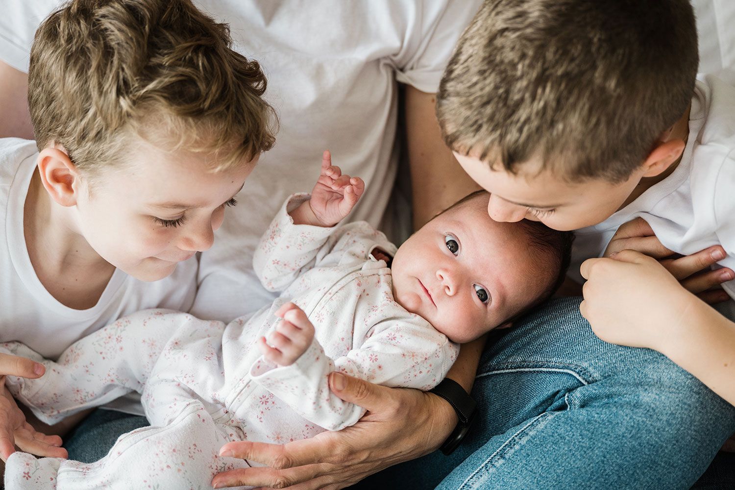 Two siblings lean in close to look at a baby lying between them wearing white clothing.