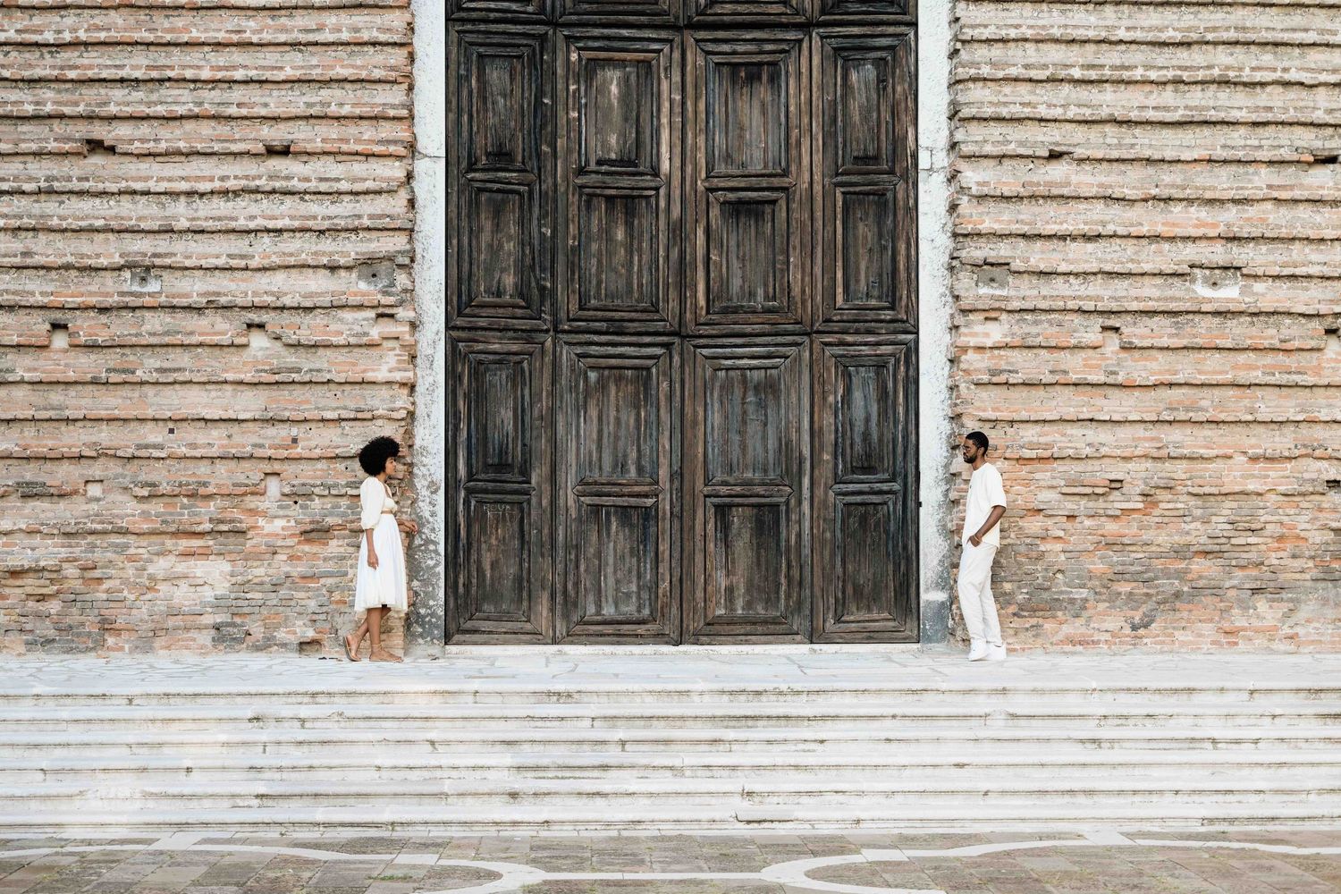 Two people in white outfits stand at opposite ends of stone steps near a large wooden door.