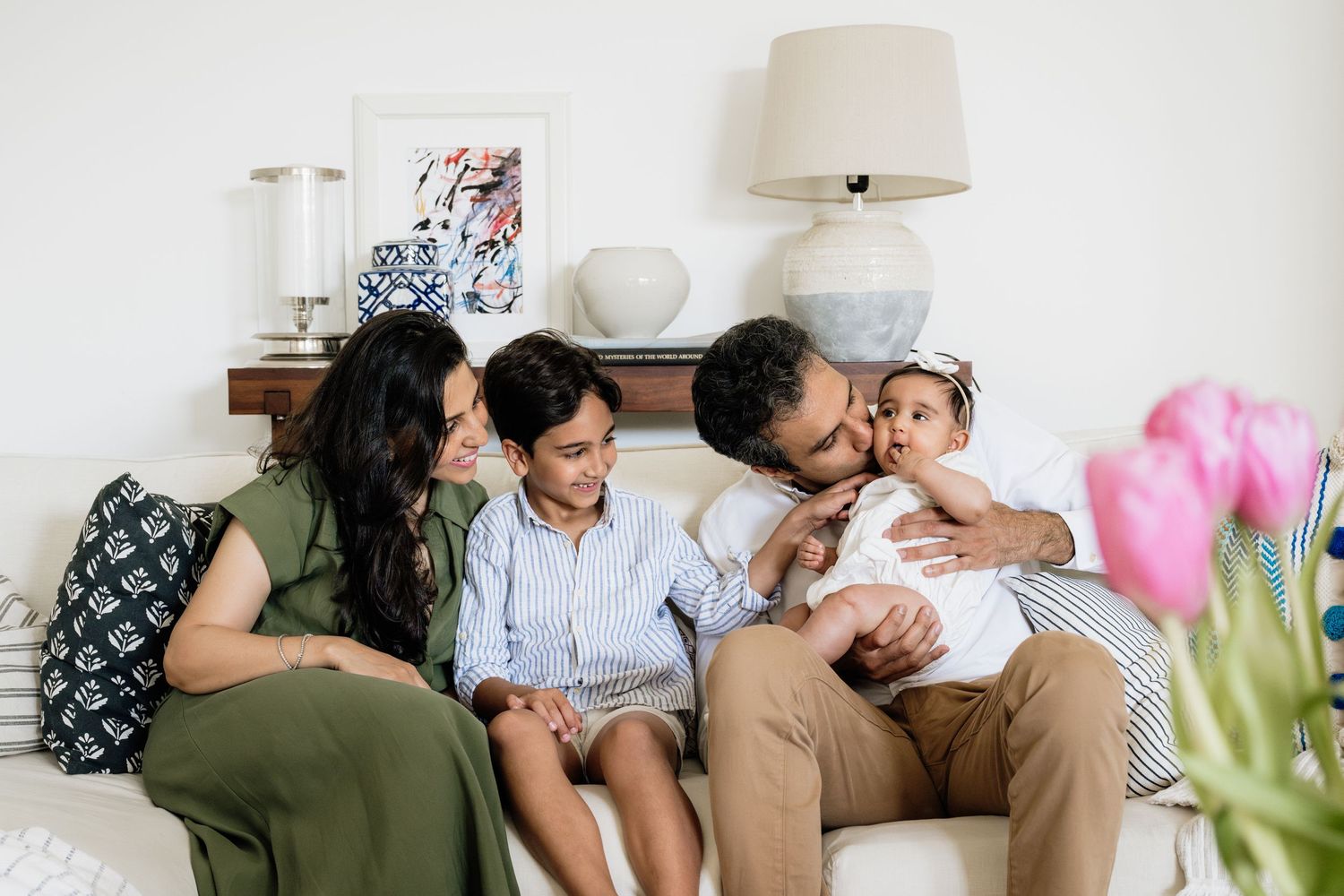 Family sharing intimate moments while lounging on white bedding in sunlit room.
