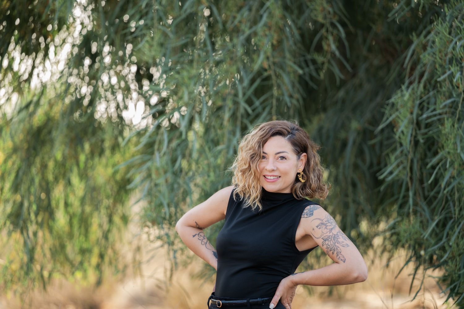 A woman poses under hanging green tree branches in a stunning outdoor setting.