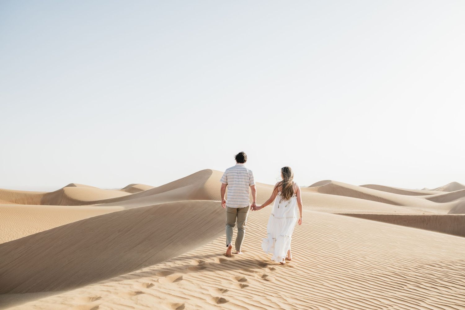 Couple walking hand in hand across vast desert sand dunes under bright sky.