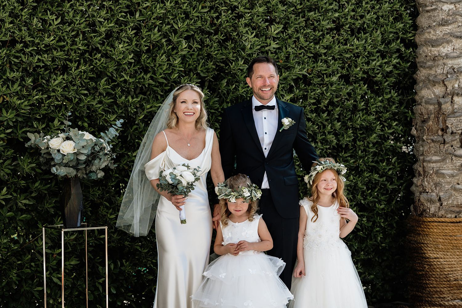 Wedding party posing against lush green hedge wall with floral decorations.