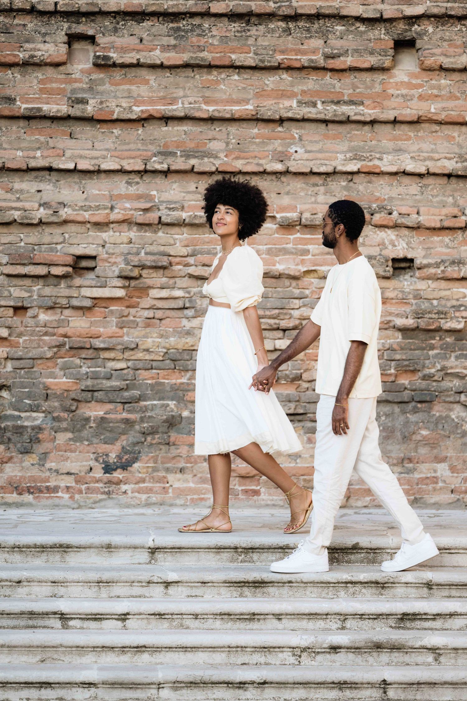 A couple in all-white outfits ascends stone steps against a rustic brick wall backdrop.