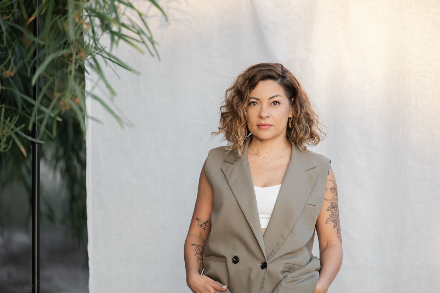 Lady wearing a sleeveless gray blazer vest poses against a white backdrop around greenery.