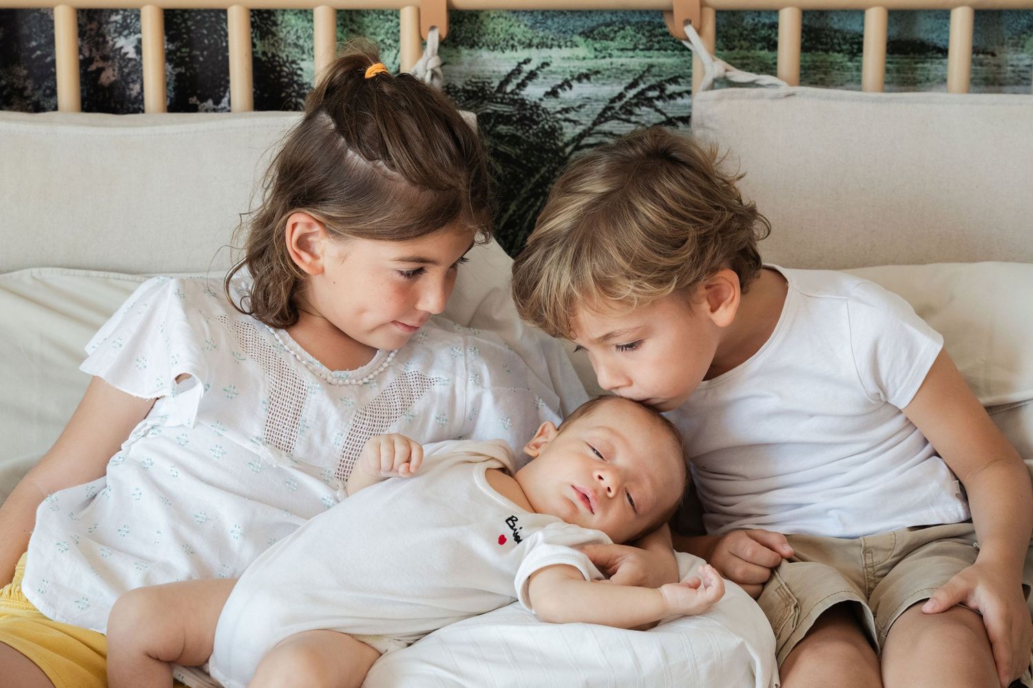 Two siblings in white shirts tenderly watching over a sleeping baby on a white bed.