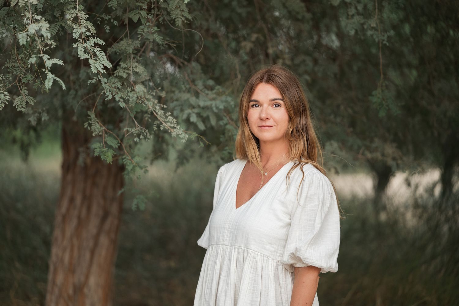 Woman in white dress poses outdoors in a natural garden setting with trees and greenery in the background.