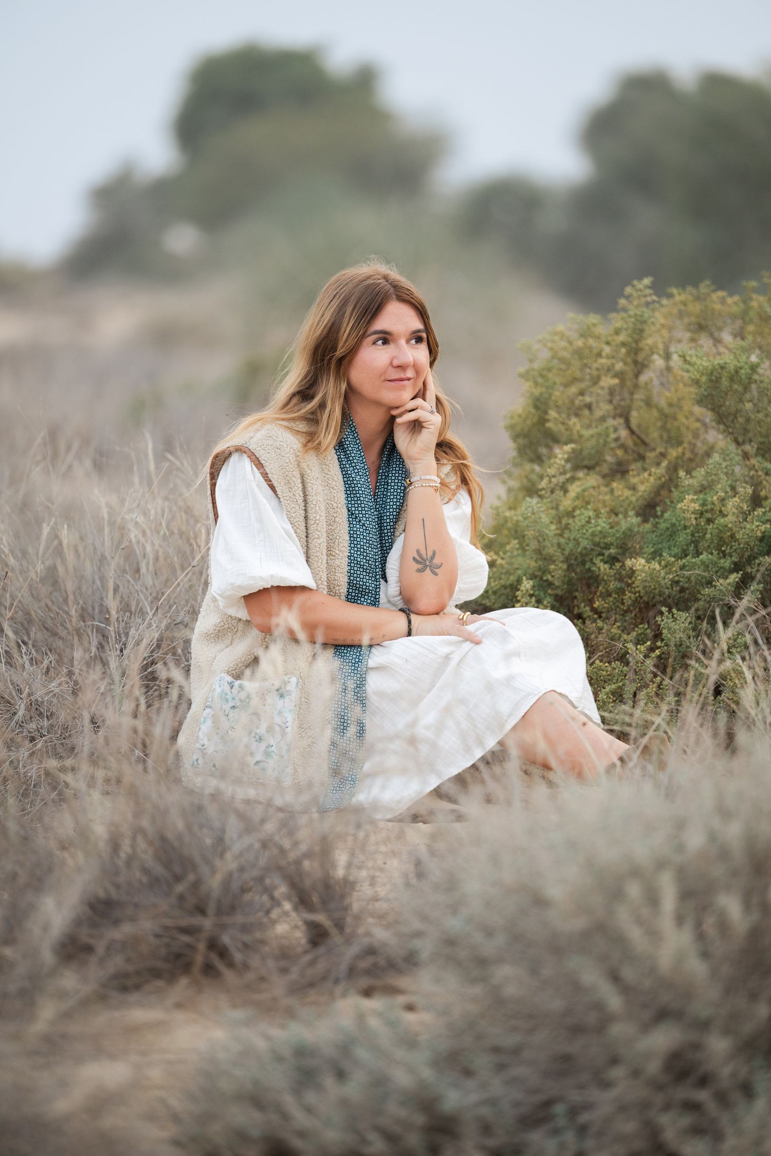 A person in a white dress and denim vest sits thoughtfully in a field of dry brush on a misty day.