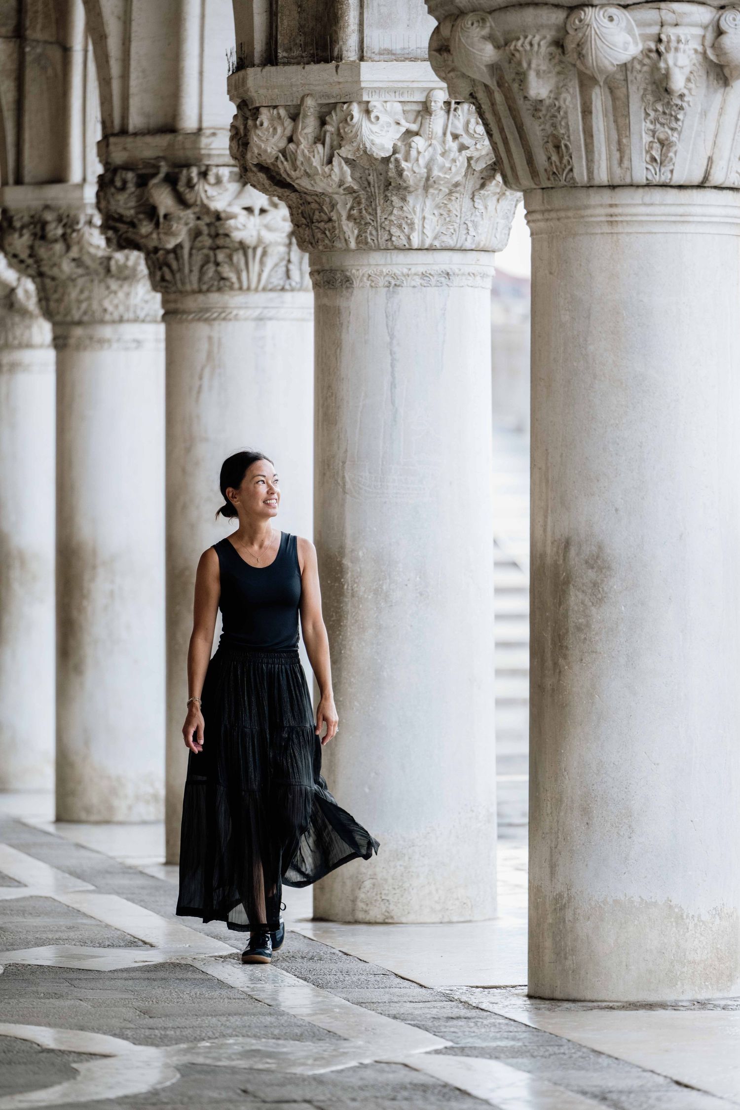 Figure in dark dress walking through historic colonnade with ornate stone columns.