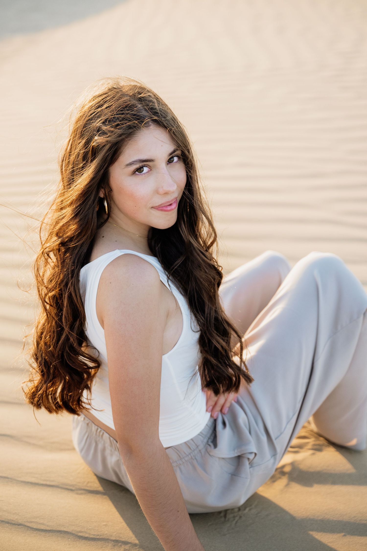 Young woman in white outfit lounging on sandy desert dunes during sunset.