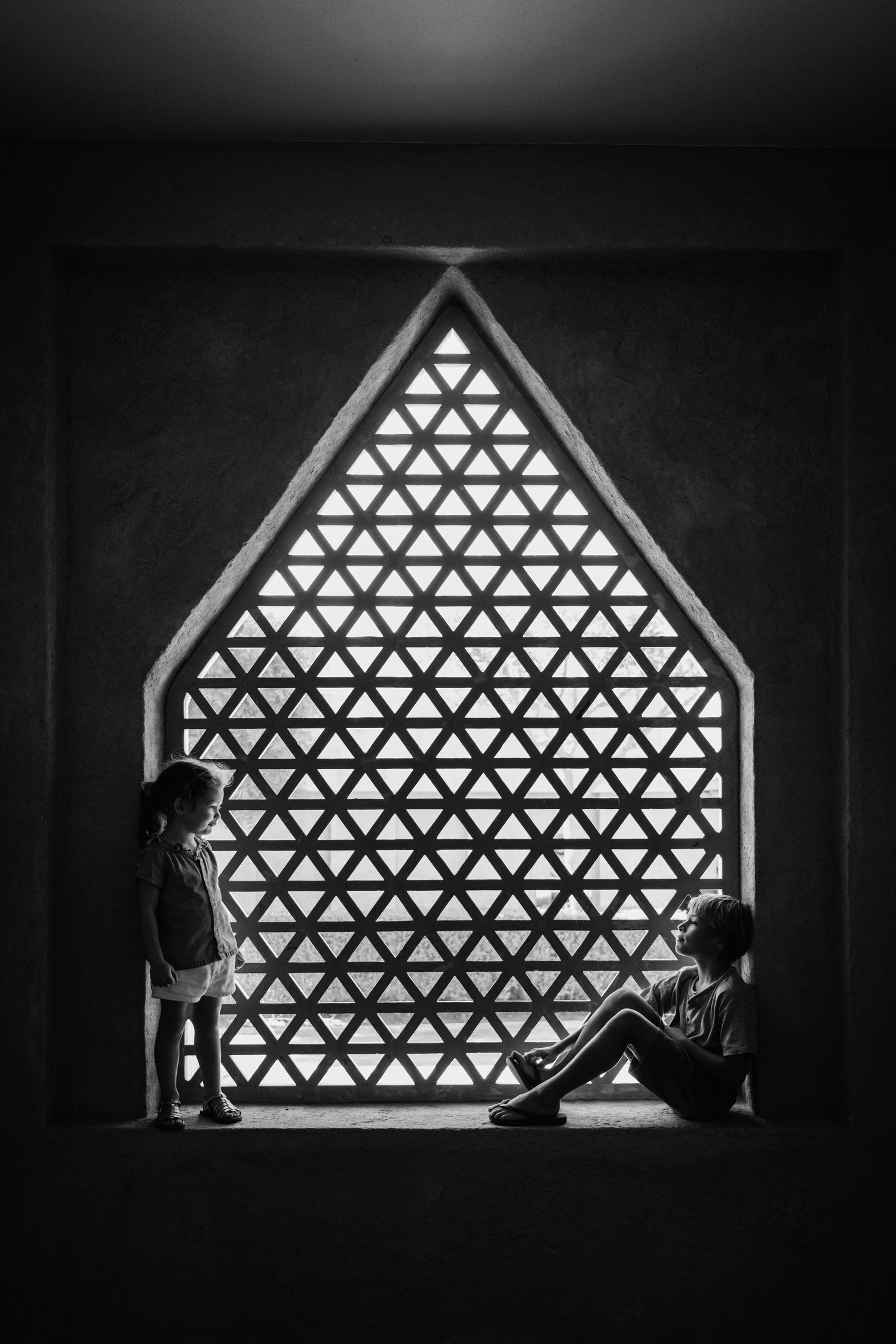 Two silhouetted figures sit by a geometric triangular arabic window with a pattern of triangles creating a striking contrast.