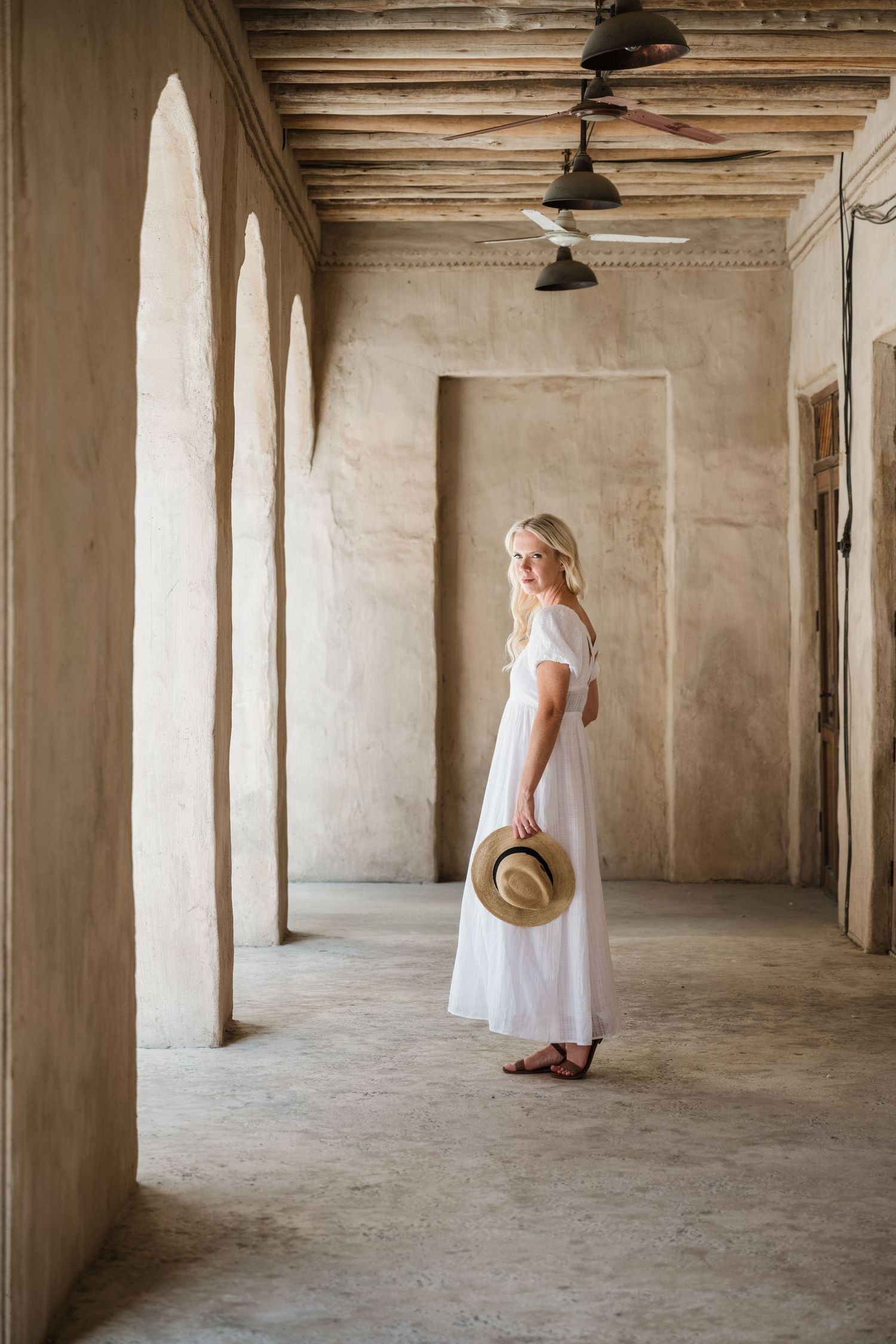 Person in flowing white dress walking through colonnade with arched pillars and dramatic lighting.