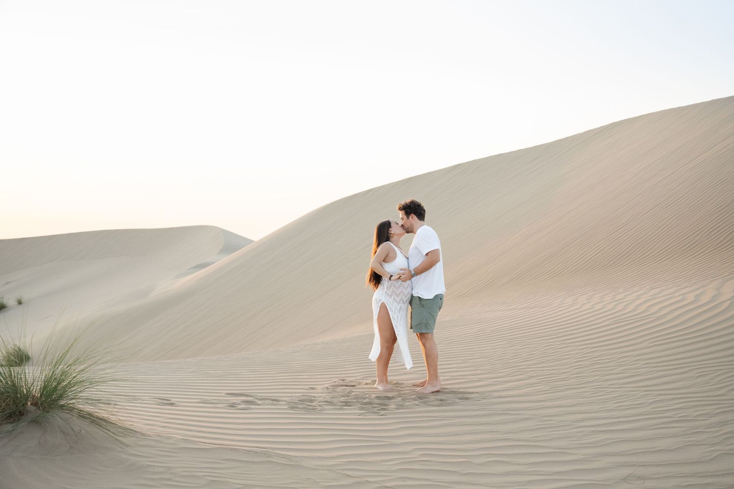 Couple in white clothing embraces on rolling sand dunes during desert photoshoot.