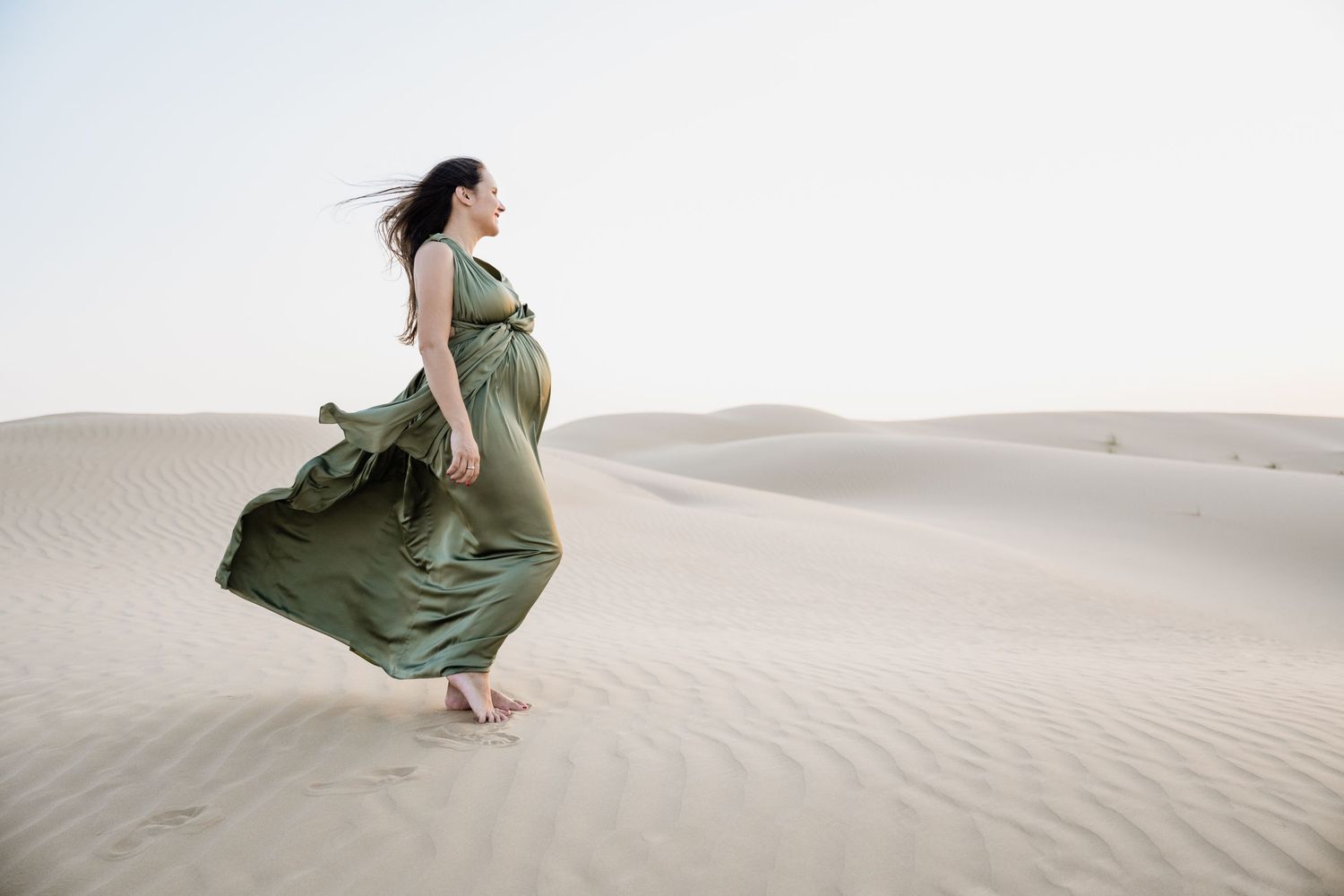 Woman in flowing green dress walks gracefully across desert sand dunes at sunset.