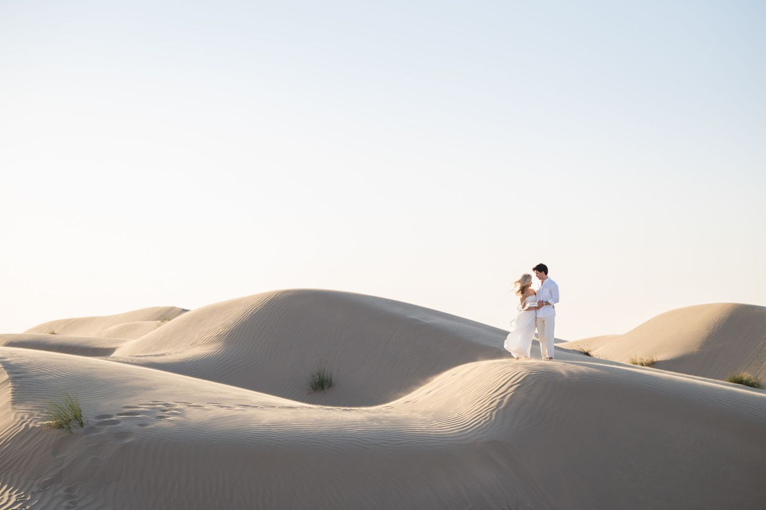 Romantic desert photoshoot with couple standing on rolling sand dunes at dusk.