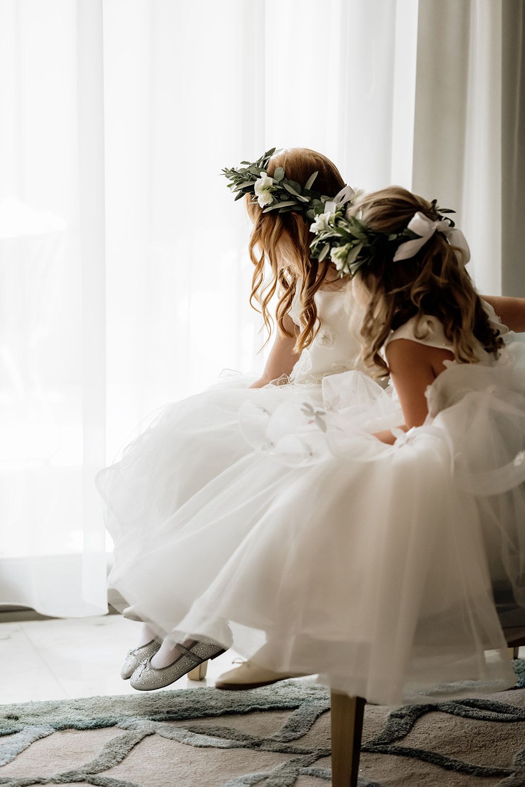 Side view of flower girls in white dresses sitting on chair against white curtains.