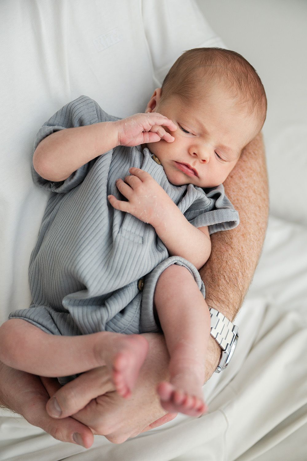 Sleeping newborn baby wearing grey onesie cradled in parent's arms on white bedding.