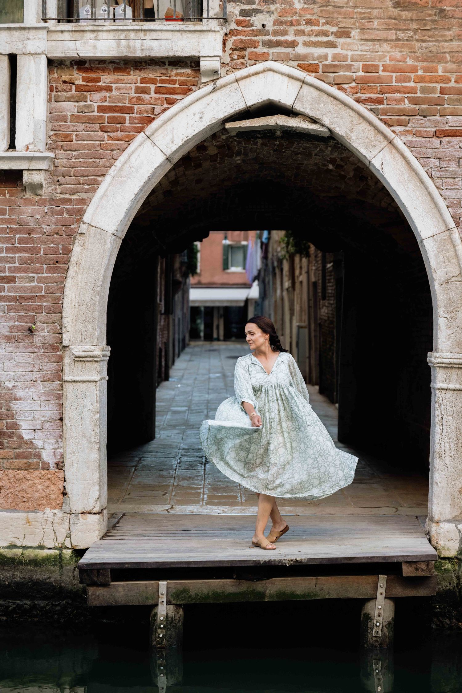 Flowing dress catches movement under brick archway along Venetian canal walkway.
