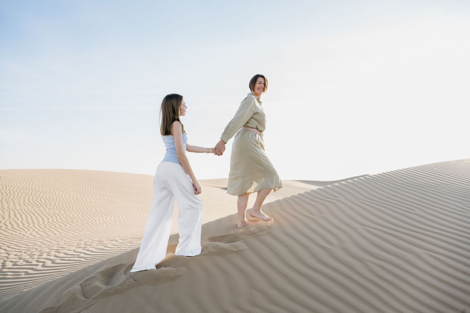 Two people climbing up steep sand dunes in light colored clothing.