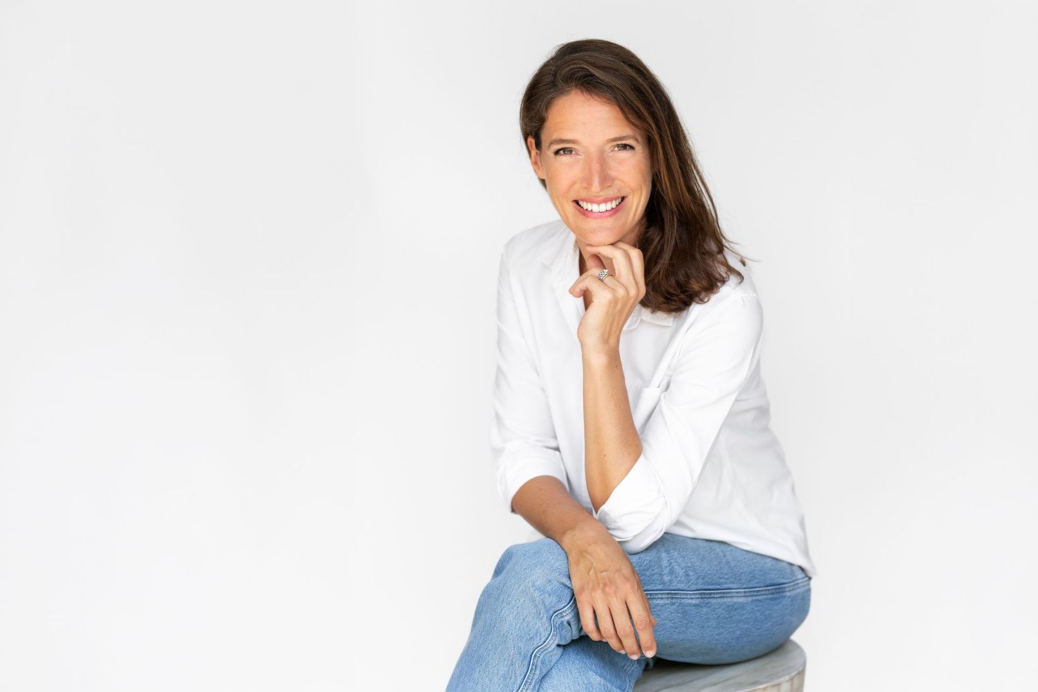 Smiling woman in white top and jeans sitting relaxed against white background with hand on chin.