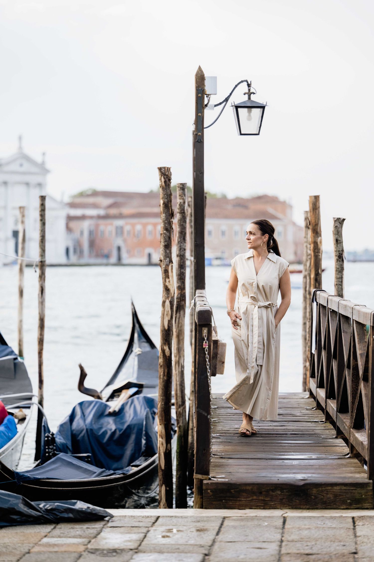 A figure in a flowing beige dress walks along a wooden dock in Venice with gondolas and historic buildings in the background.