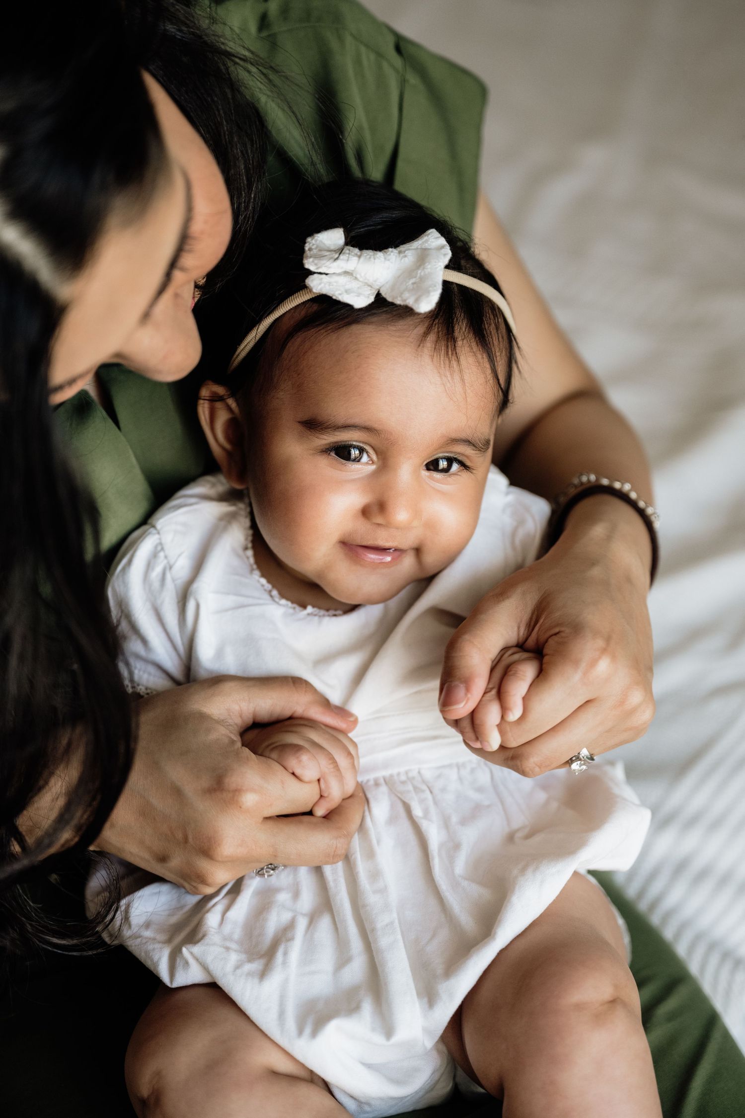 Baby in white outfit with bow smiles during intimate photo session with parent.