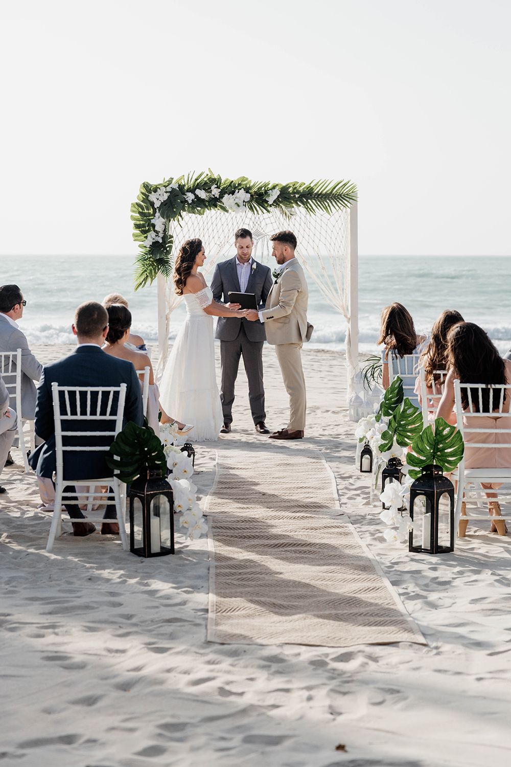A beach wedding ceremony setup with white chairs, lanterns, and tropical greenery along a sandy aisle.