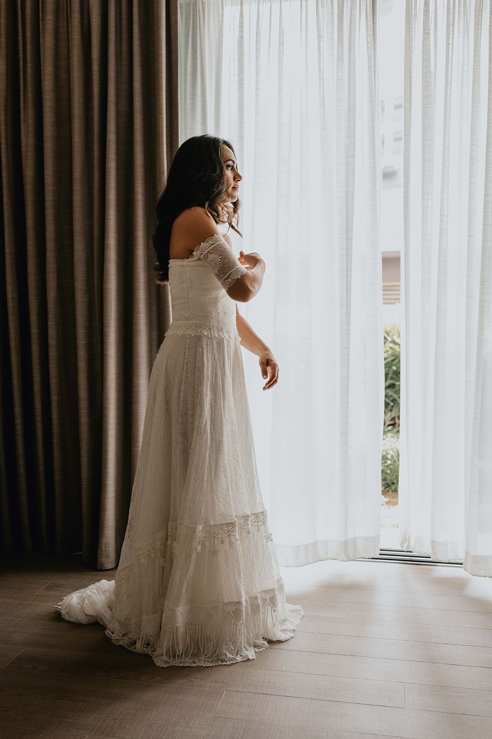 Bride in flowing lace gown standing by window with sheer curtains.