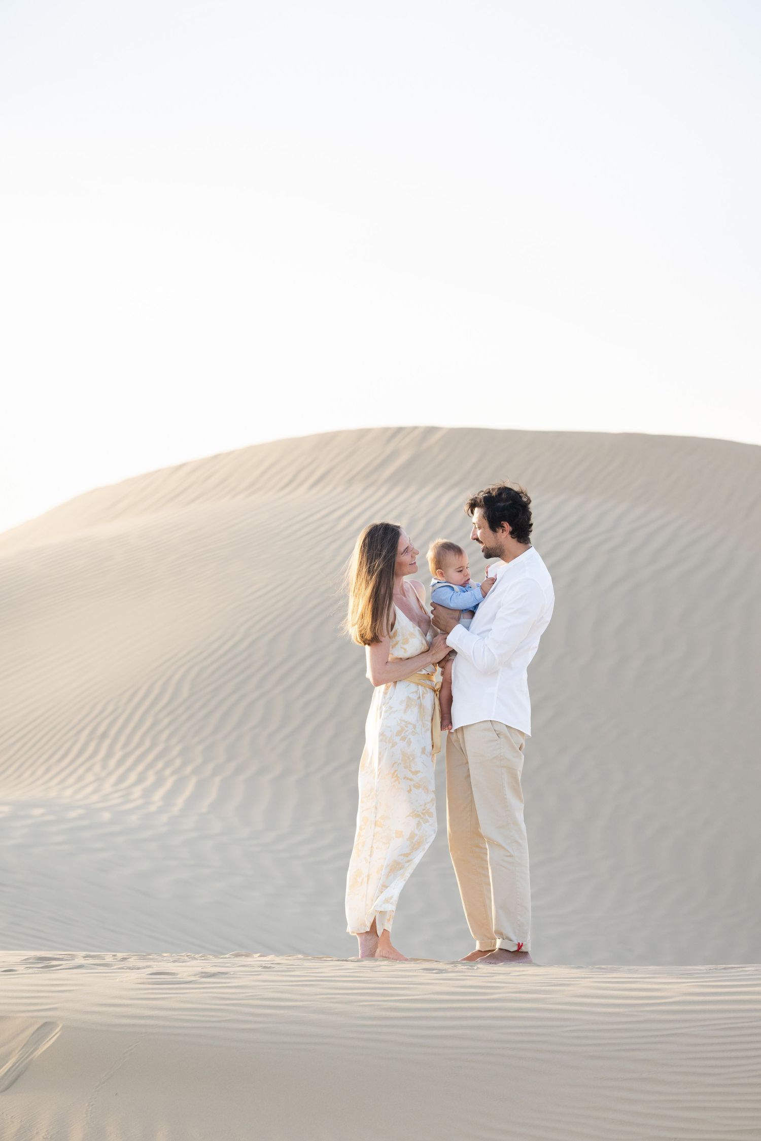 A couple in light-colored clothing poses together on sandy desert dunes at sunset.