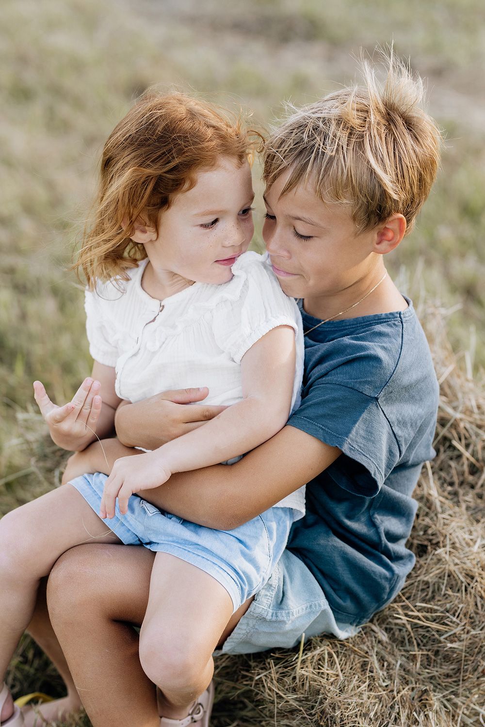 A series of tender moments captured between two siblings sitting in a grassy field.