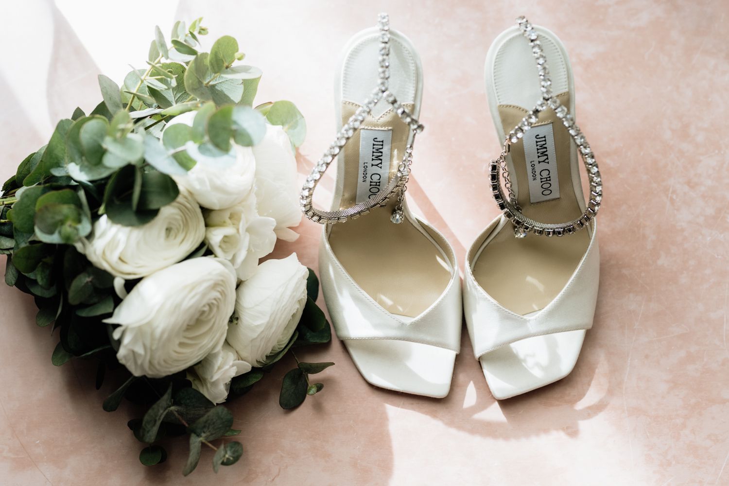 Wedding accessories including white roses and pearl-adorned bridal shoes on pink background.