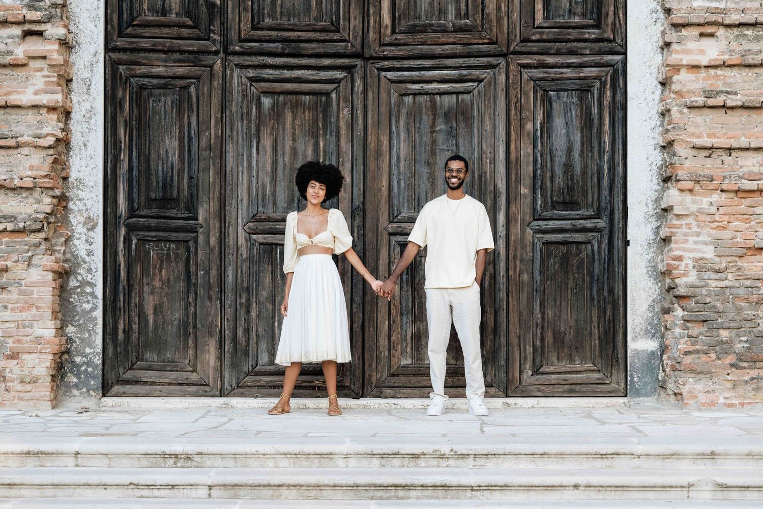 A couple in white outfits poses holding hands on stone steps against a rustic door.