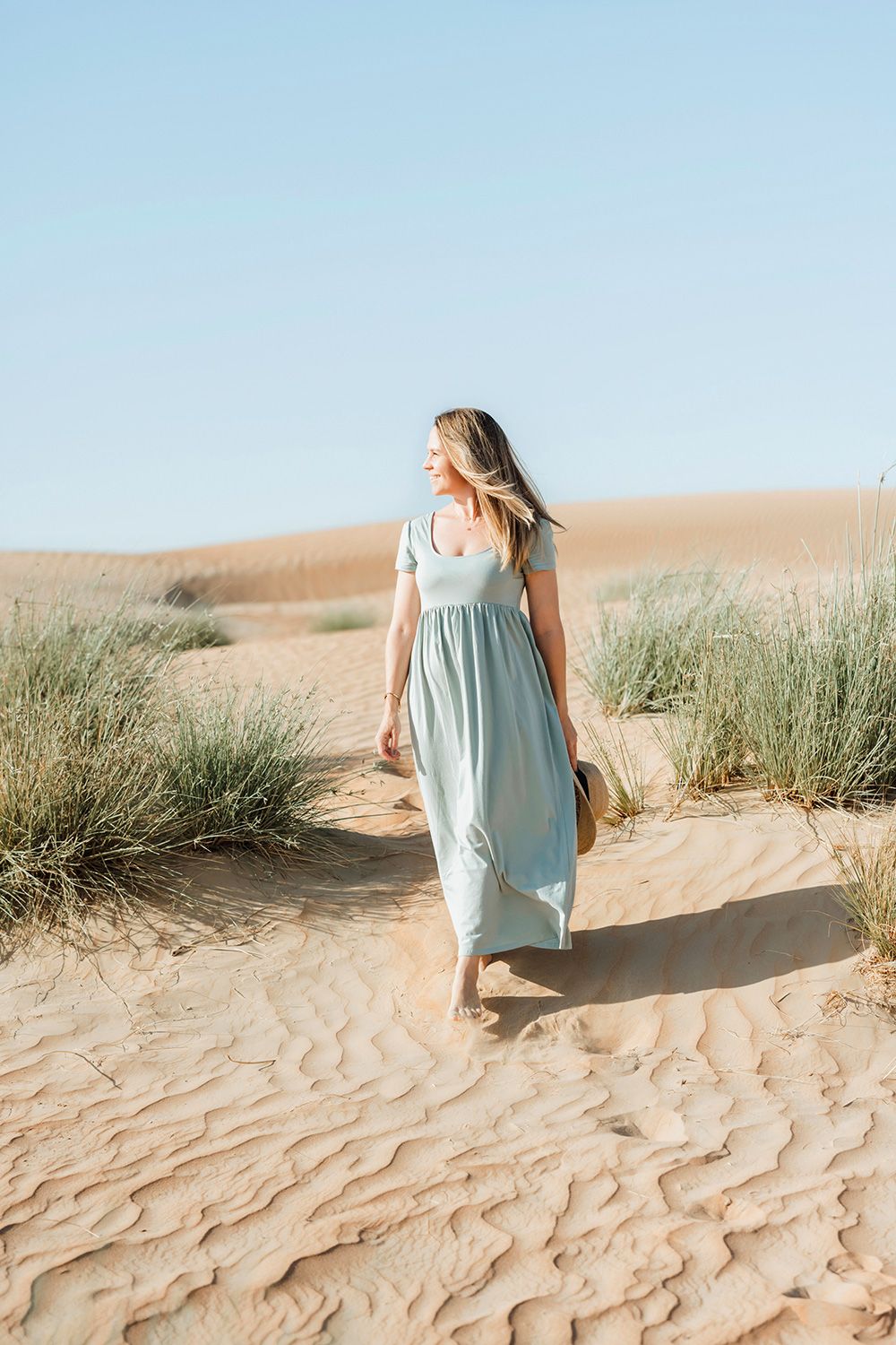 Woman in flowing light blue maxi dress walks through sandy dunes with beach grass on a sunny day.