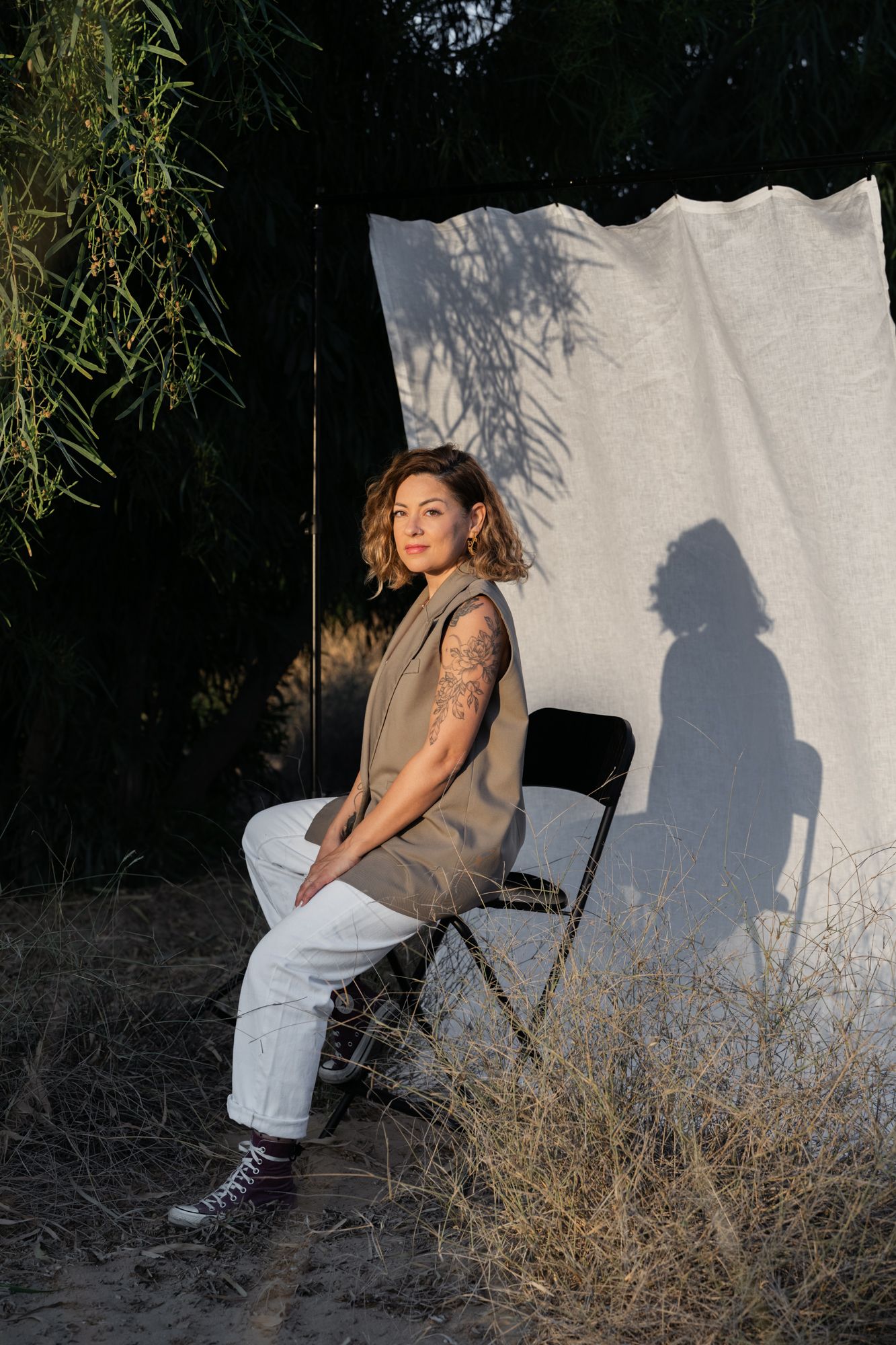 A woman in casual attire sits on a folding chair against a white backdrop with leaves and shadow patterns.