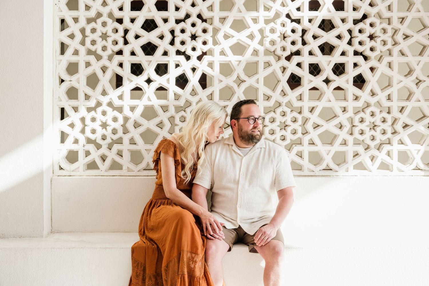 Couple sitting together against decorative white lattice wall during intimate photoshoot.