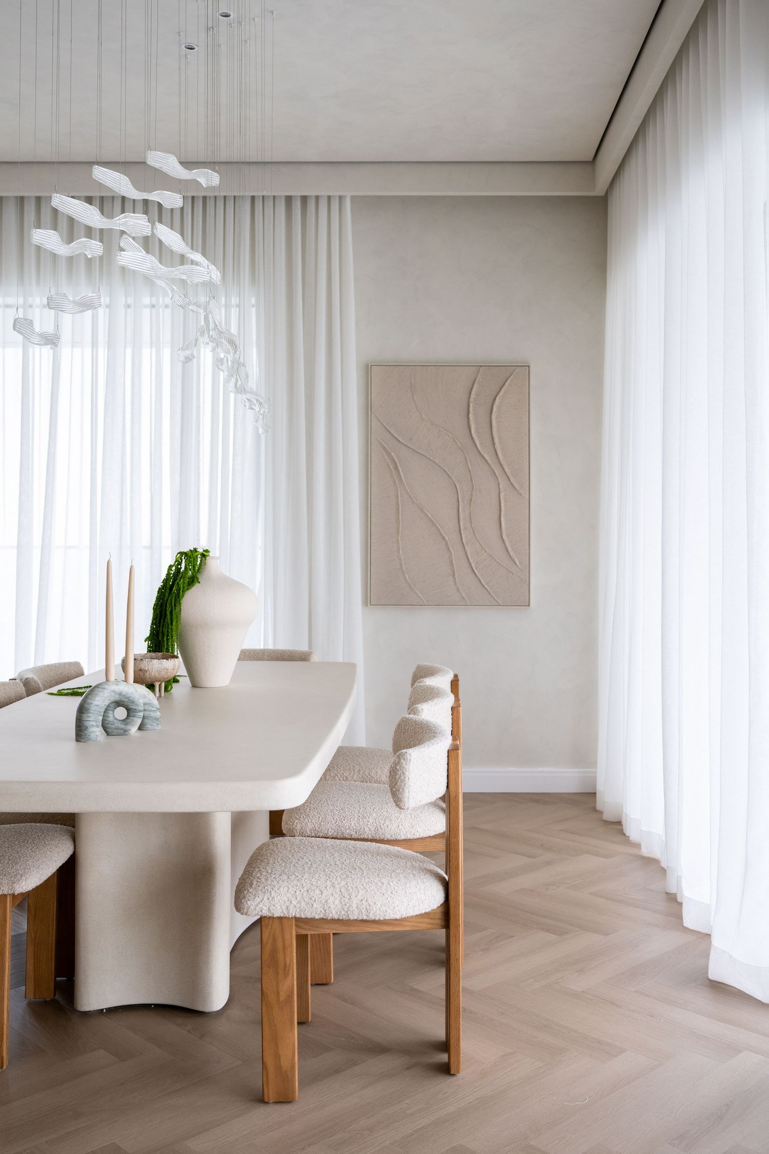 Minimalist dining room with cream bouclé chairs, white sculptural table, herringbone floors, sheer curtains, and abstract wall art.