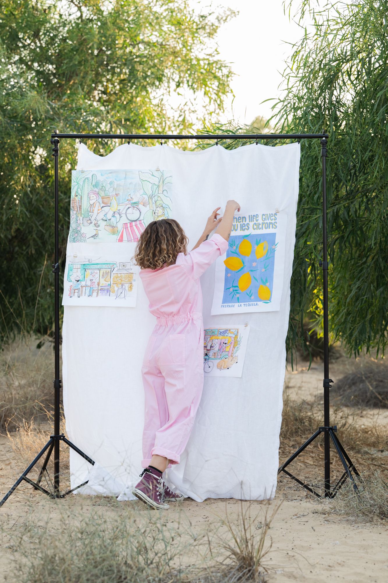 Artist in pink jumpsuit hangs artwork on white backdrop display stand outdoors among green trees.