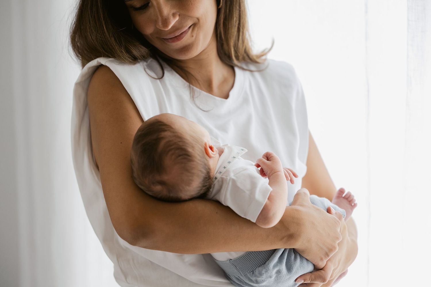 Tender moment between parent and newborn baby in white clothing by window light.