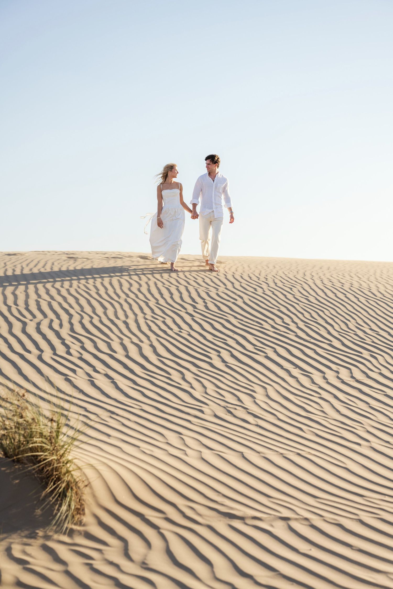 A couple in white attire walking along rippling sand dunes during a golden desert sunset.