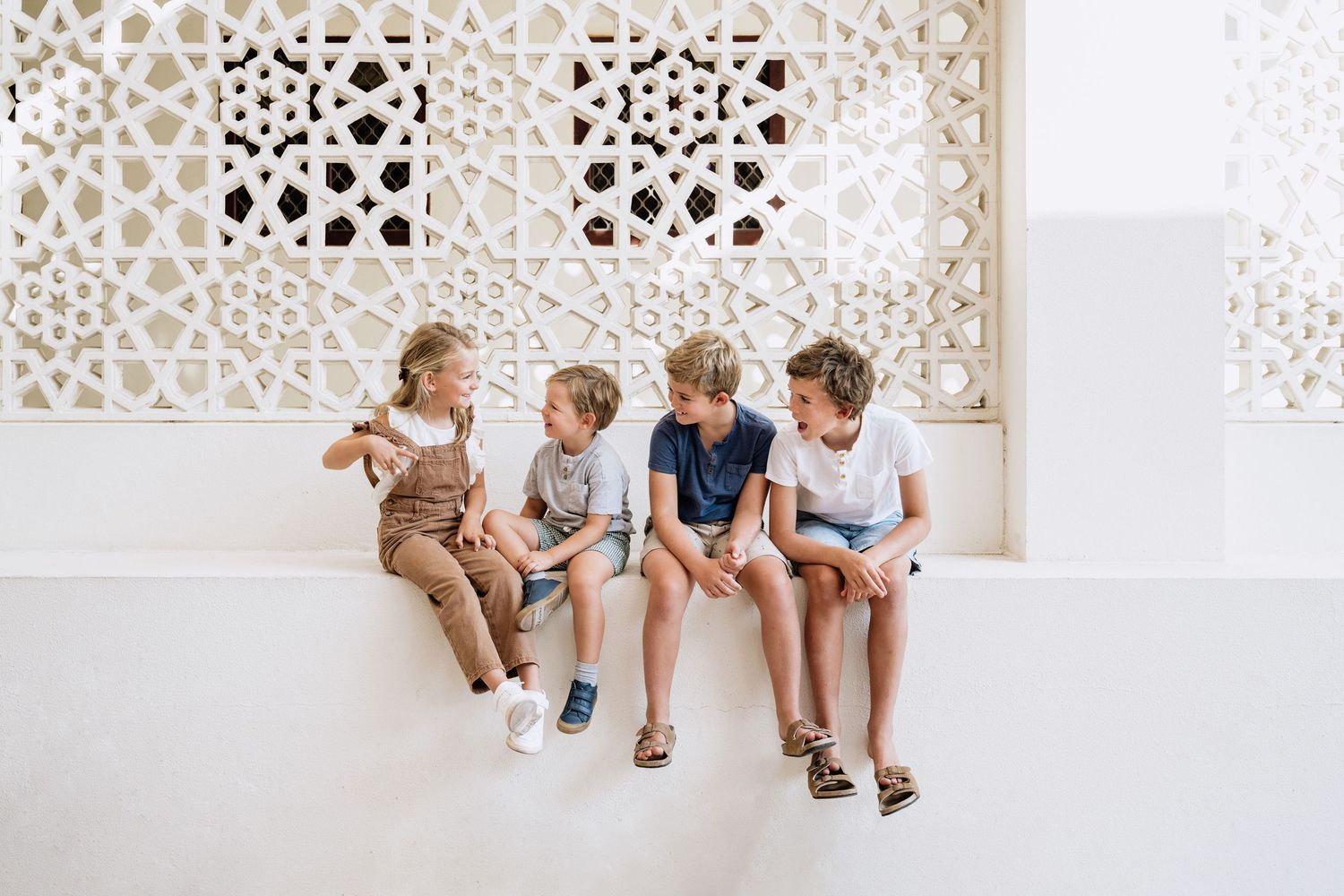 Four children sit together on a white bench against a decorative wall with geometric patterns.