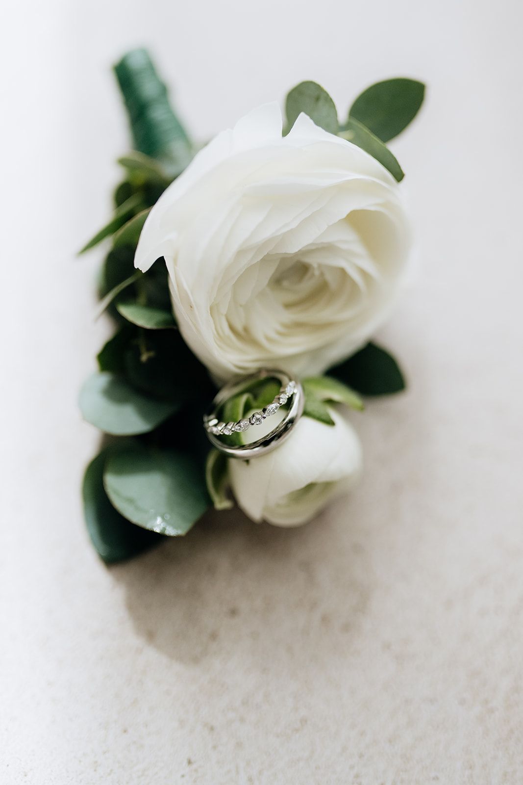 White rose boutonniere with green leaves and wedding ring detail on light surface.