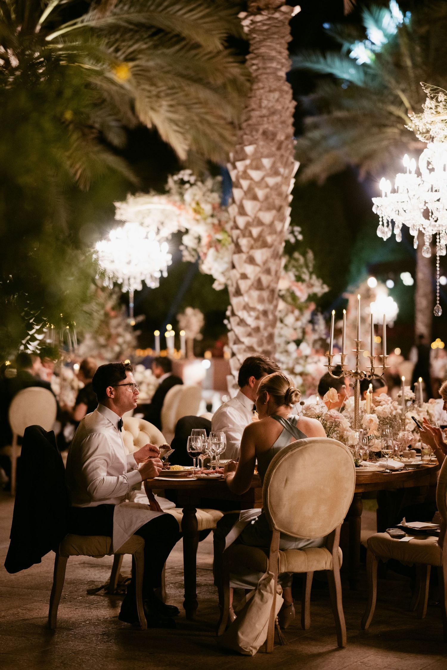Romantic outdoor wedding reception with candlelit tables under palm trees at night.