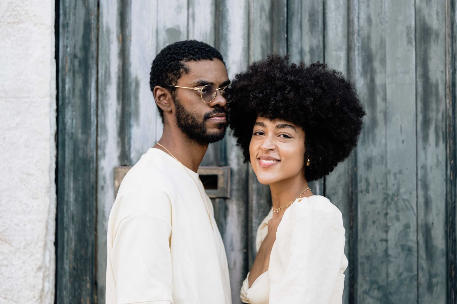 Happy couple in coordinated white outfits posing against rustic backdrop.