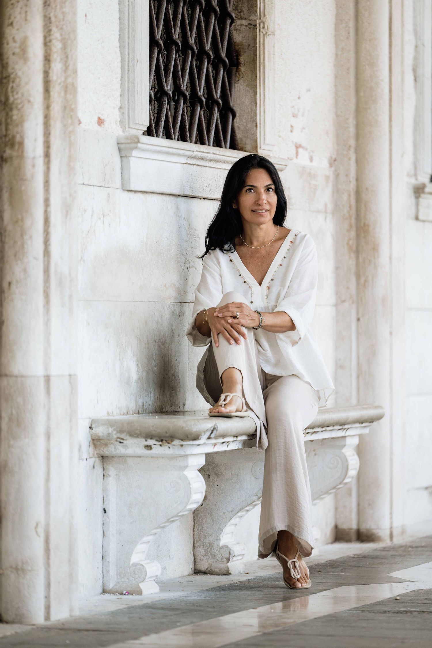 Figure in white dress seated among classical white columns and arches.